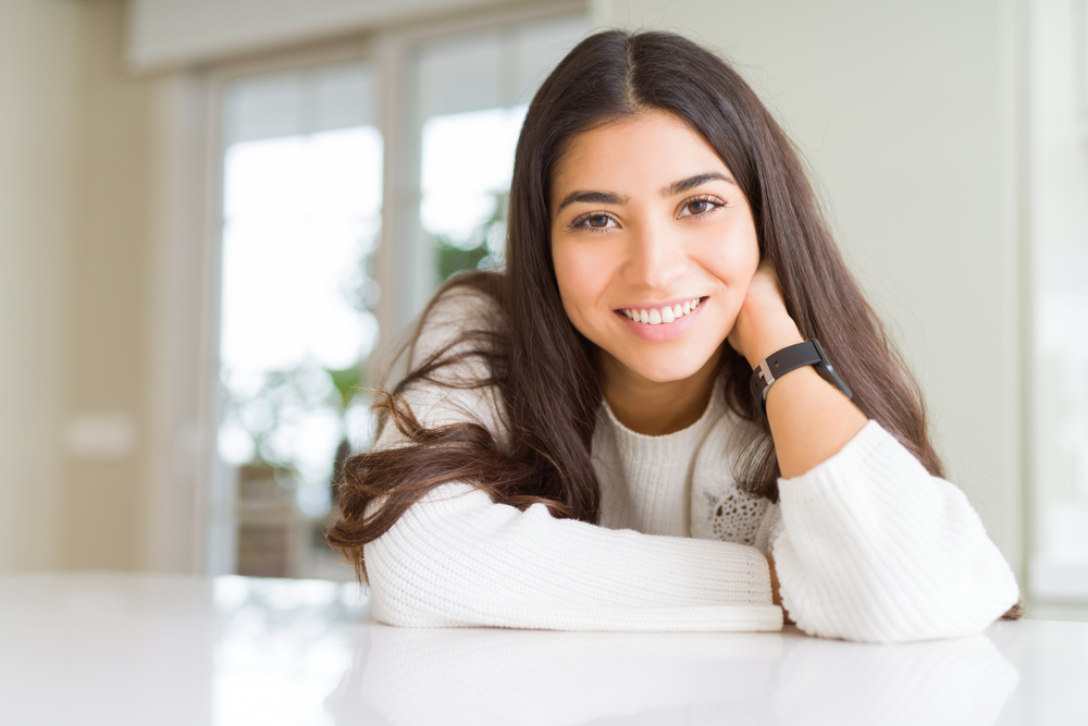 Confident woman with a healthy smile at home – Cleaning and Dental Exam A relaxed young woman in a white sweater smiles naturally while resting her chin on her hand, reflecting the long-lasting benefits of professional oral hygiene – Cleaning and Dental Exam.