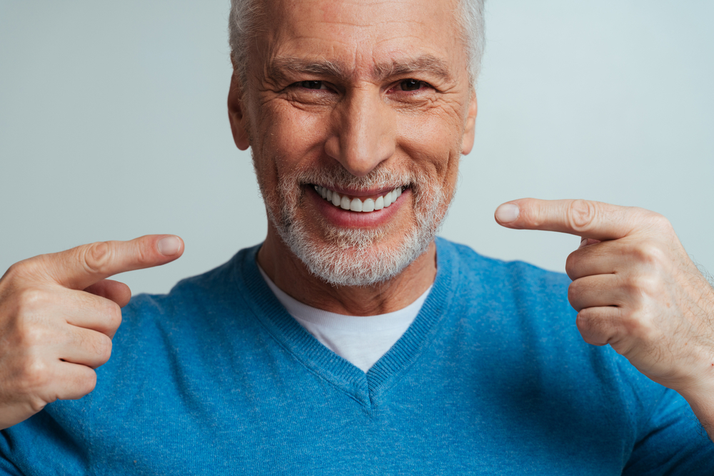 Mature man showing off healthy white teeth – Teeth Cleaning in Arlington A cheerful man in a blue sweater proudly points to his bright smile, highlighting the benefits of regular dental cleanings for long-term oral health – Teeth Cleaning in Arlington.