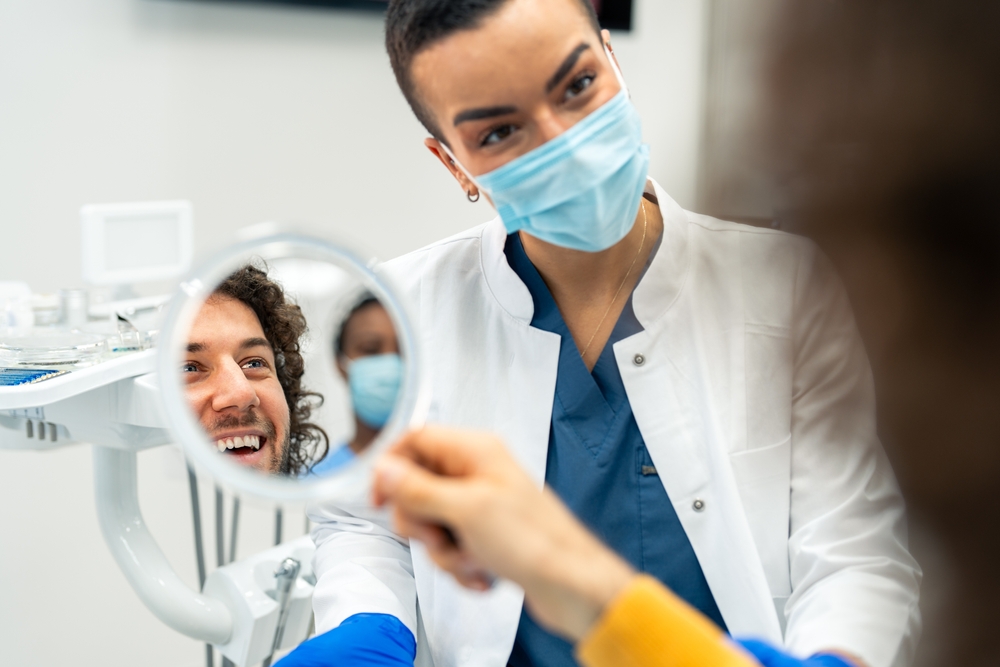 A happy patient looks into a handheld mirror while the dentist smiles behind a mask, reflecting satisfaction after a successful treatment – Dental Fillings.