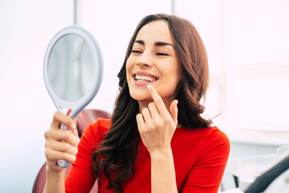 Woman admiring her smile in the mirror – Cleaning and Dental Exam A happy dental patient wearing a red top looks at her reflection and points to her bright smile after a successful appointment. The image highlights satisfaction and confidence in dental results – Cleaning and Dental Exam.