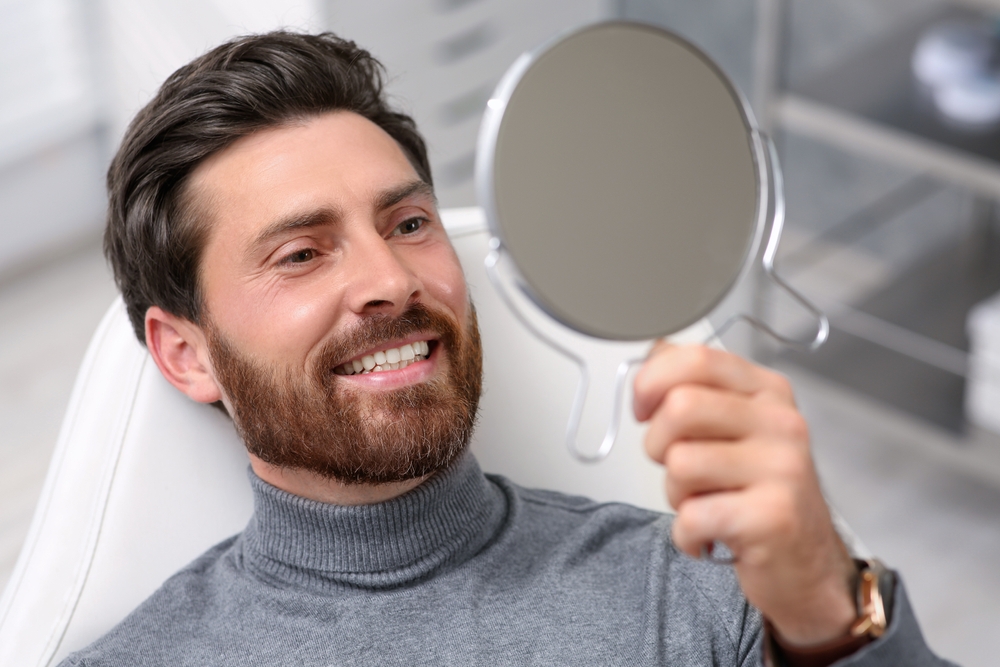 Patient admiring restored smile in mirror – Dental Implants in Arlington A man with a beard smiles confidently while holding a mirror after his implant procedure, showcasing his bright, natural-looking teeth – Dental Implants in Arlington.