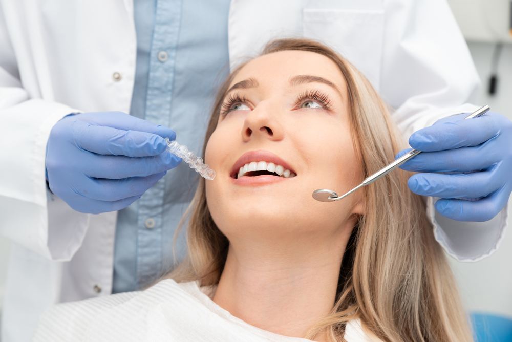 A dental professional wearing blue gloves holds a clear aligner while a woman in the chair smiles confidently during her appointment – Dental Fillings.