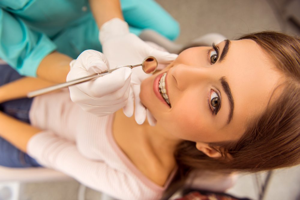 A young woman with braces smiles during a dental examination as the dentist inspects her teeth with a mirror tool, highlighting professional orthodontic care – Emergency Dentistry.