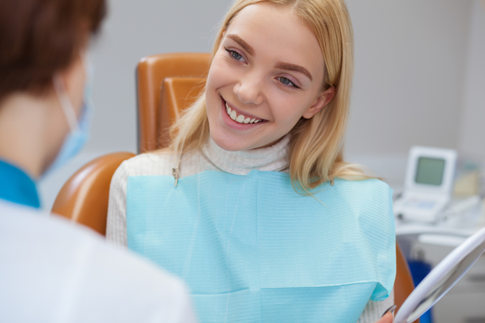 Smiling patient during dental consultation – Cleaning and Dental Exam A cheerful woman sits in the dental chair, smiling at her dentist during a routine checkup. The scene captures a comfortable, friendly atmosphere of professional oral care – Cleaning and Dental Exam.