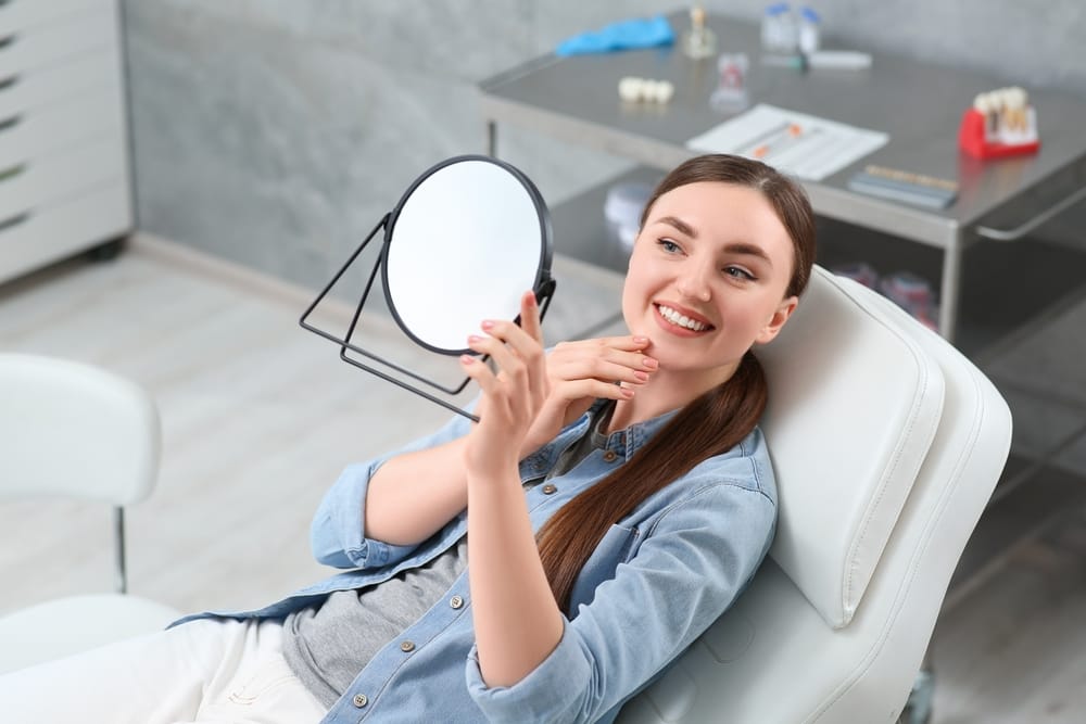 A smiling woman holds a mirror and admires her bright teeth after treatment, reflecting confidence and satisfaction with expert dental care – Emergency Dentistry.