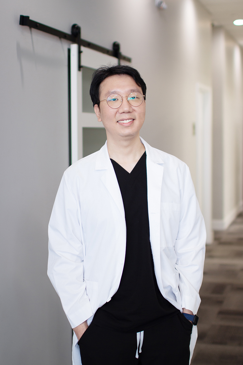 Dr. Dongho Kim, DDS professional portrait in dental office hallway – Dentist Arlington WA Smiling Dr. Dongho Kim, DDS, wearing glasses and a white coat while standing in a bright dental office hallway – Dentist Arlington WA