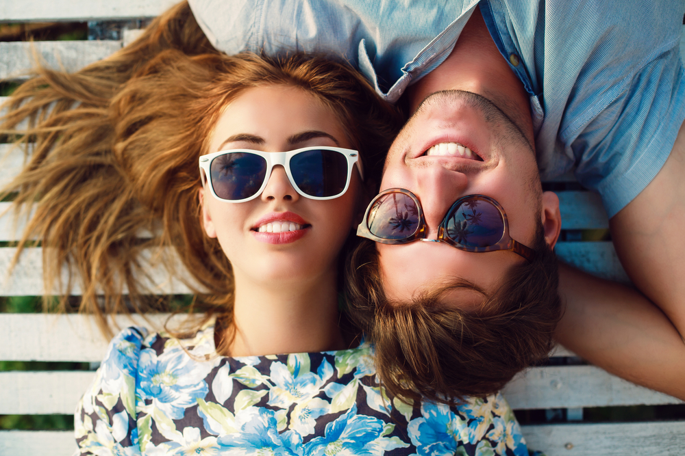 A young couple wearing sunglasses lies side by side outdoors, showing off their radiant, healthy smiles under the sunlight – Dental Fillings.