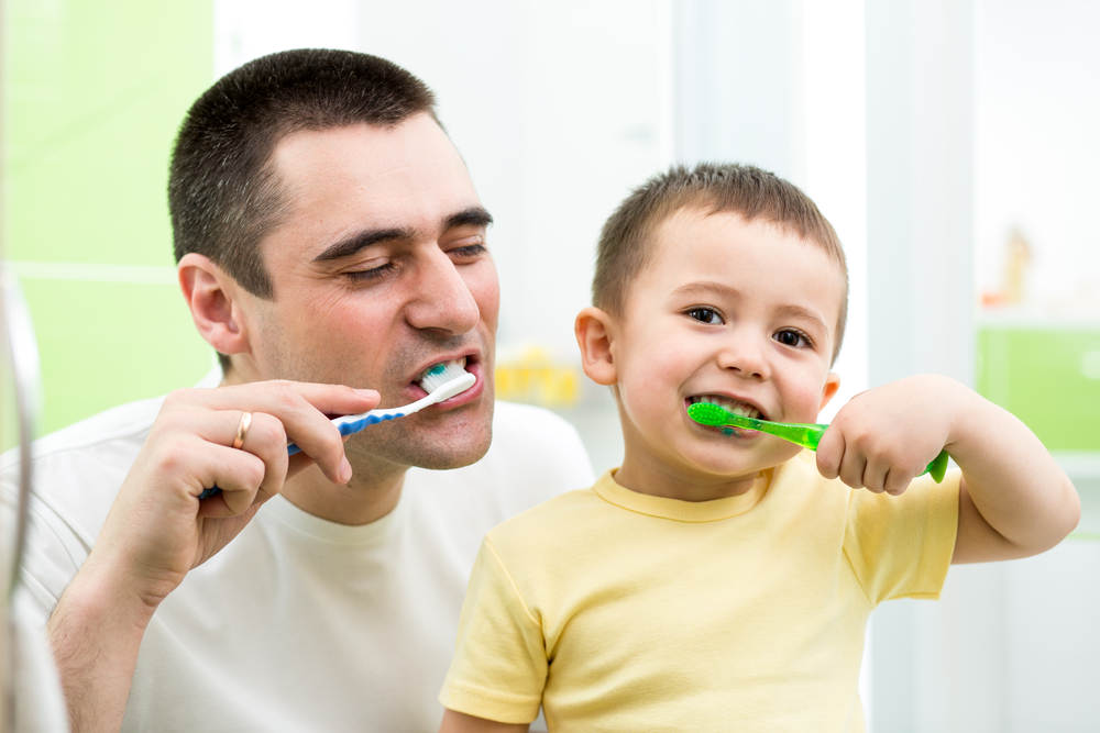 Father and young son brushing teeth together – Dentist Arlington WA Smiling father and little boy brushing their teeth side by side in a bright bathroom, promoting healthy dental habits – Dentist Arlington WA