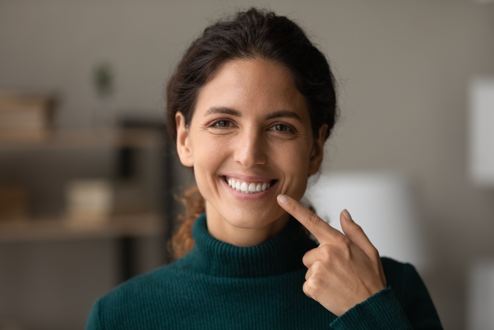 Woman pointing to her clean, bright smile – Teeth Cleaning in Arlington A smiling woman in a green sweater points to her freshly cleaned teeth, showcasing confidence and satisfaction after a professional dental cleaning – Teeth Cleaning in Arlington.